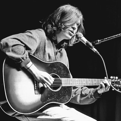 Black and white photo of young man with shoulder-length hair singing on stage with an acoustic guitar