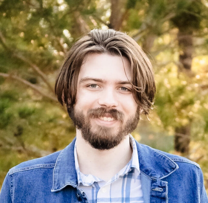 Young man with brown hair and jean jacket smiles at camera